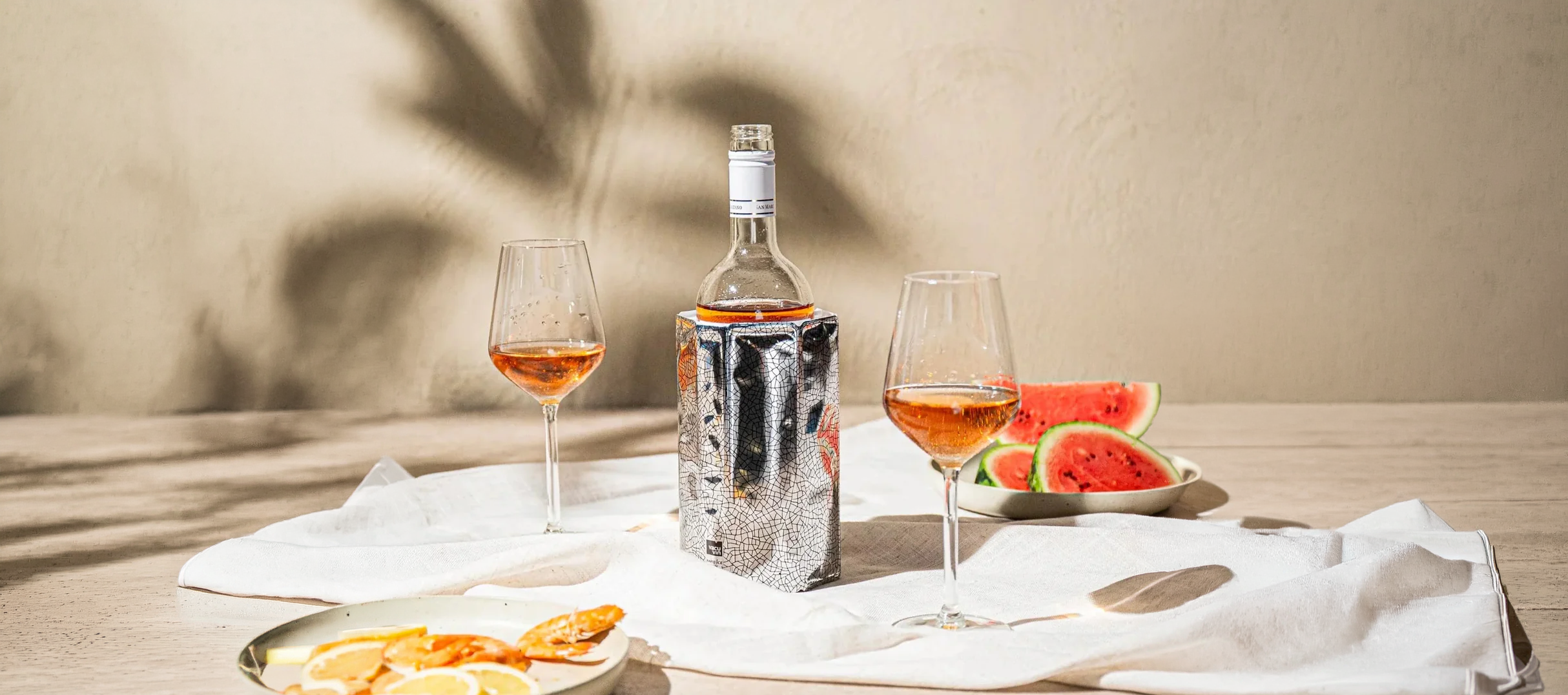 Wine bottle with two glasses of wine, a plate of snacks, and watermelon slices on a wooden table.